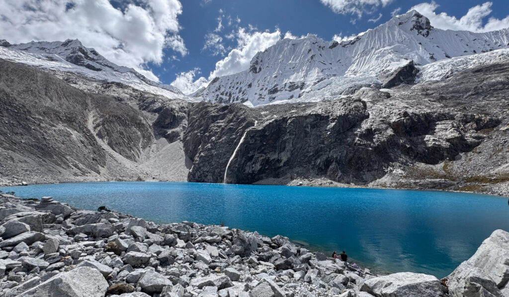Un lac glaciaire bleu vif, qui fait partie du célèbre Trek du Huayhuash, est entouré d'un terrain rocheux et de montagnes enneigées sous un ciel partiellement nuageux. Deux personnes sont assises parmi les rochers près du bord de l'eau au premier plan. - Karavaniers