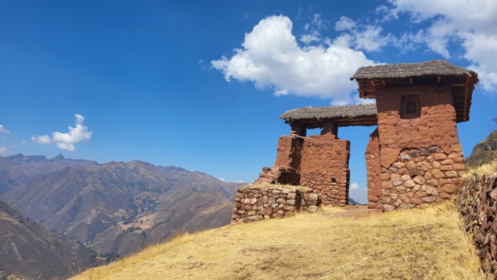 Une porte en pierre et en adobe avec un toit de chaume se dresse sur une colline sèche et herbeuse en Amazonie au Machu Picchu, surplombant un paysage montagneux sous un ciel bleu avec des nuages épars. - Karavaniers