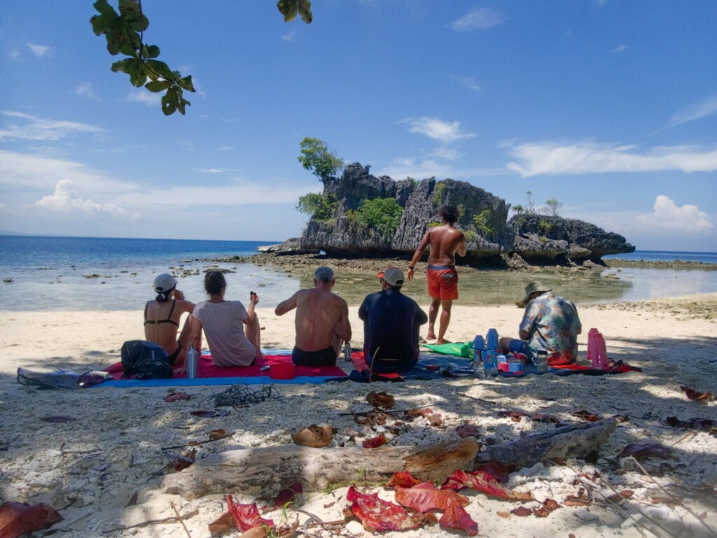 Groupe assis sur le sable partageant un repas sous les arbres, en bord de mer