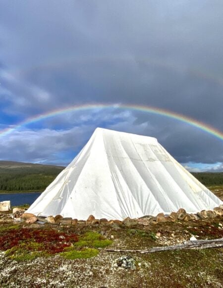 Une tente blanche sur fond de toundra, traversée par un arc-en-ciel