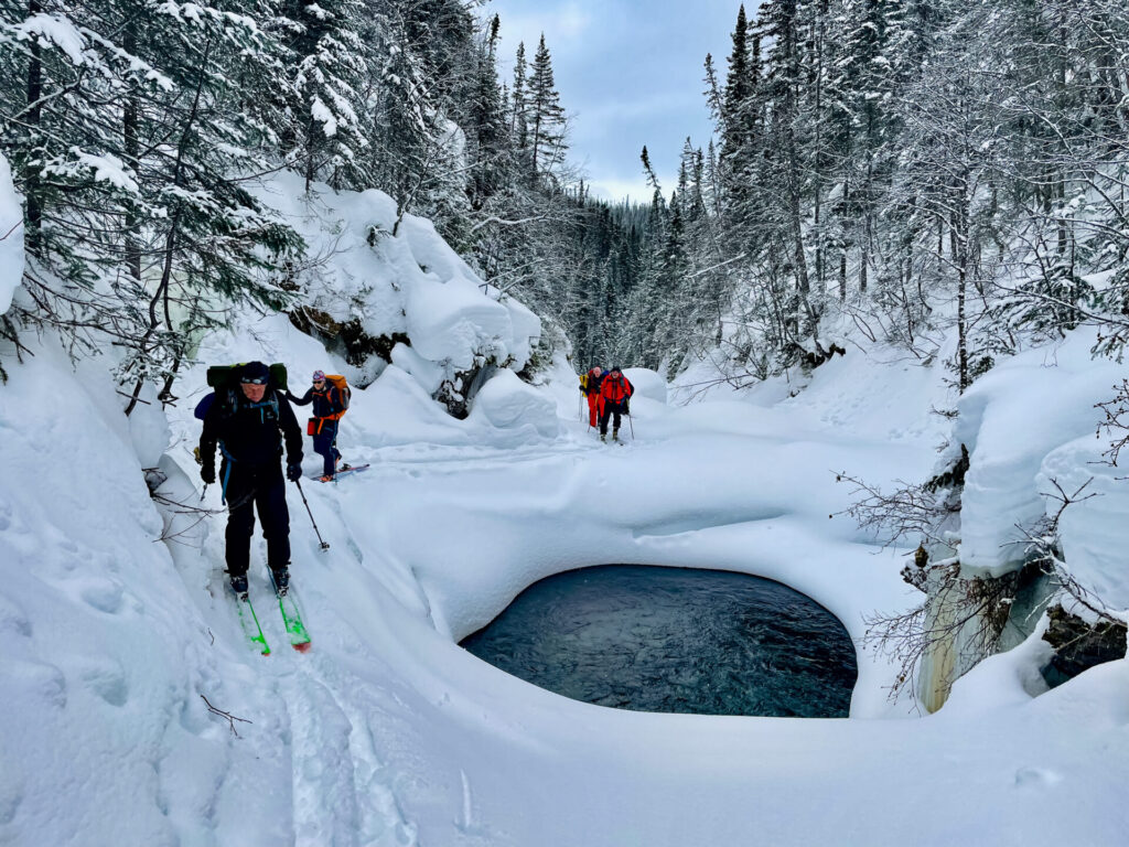 Quatre personnes en tenue d'hiver et en skis traversent le paysage enneigé et boisé des monts Groulx de l'Uapishka, en suivant une rivière partiellement gelée avec un bassin non gelé, entouré d'arbres et de falaises enneigés. - Karavaniers