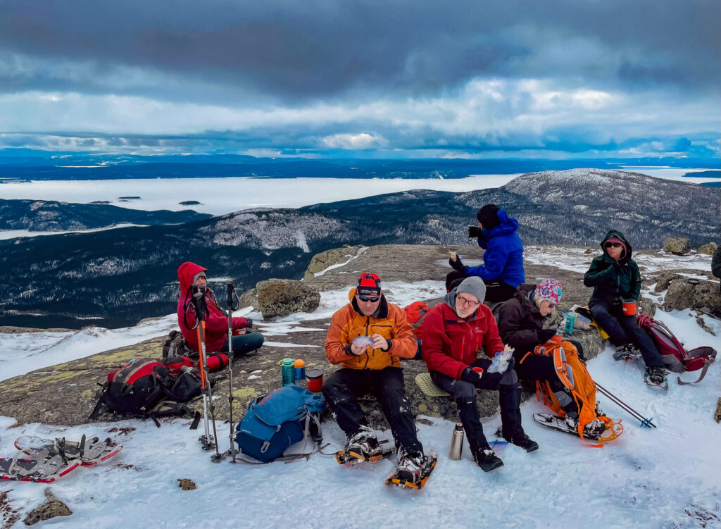Un groupe de six personnes portant des vêtements d'hiver est assis sur le sommet enneigé du mont Groulx à Uapishka, entouré de sacs à dos et de bâtons de trekking, avec une vue panoramique sur les montagnes, un lac gelé et un ciel nuageux - un endroit idéal pour les aventures à ski. - Karavaniers