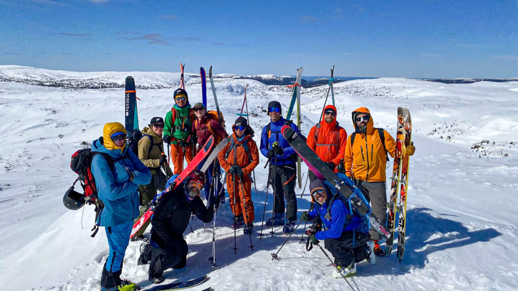 Un groupe de onze personnes en tenue d'hiver colorée se tient debout et à genoux avec des skis et des bâtons sur les pentes enneigées des monts Groulx de l'Uapishka, sous un ciel bleu clair. - Karavaniers