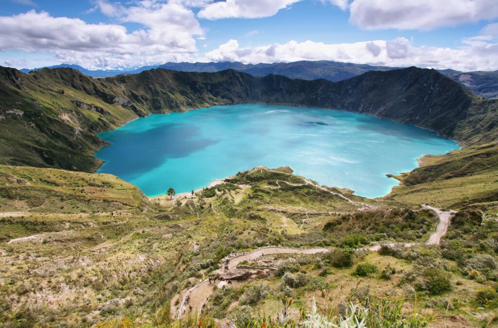 Un lac de cratère turquoise est entouré de montagnes vertes escarpées sous un ciel partiellement nuageux. Des sentiers sinueux le long de l'avenue des Volcans et une végétation clairsemée au premier plan descendent vers l'eau. - Karavaniers