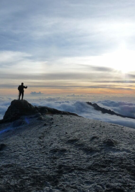 Une personne se tient sur un sommet rocheux couvert de givre au lever du soleil, pointant vers l'horizon avec des nuages en dessous et les sommets lointains du Kilimandjaro et du Meru visibles sous un ciel partiellement nuageux. - Karavaniers