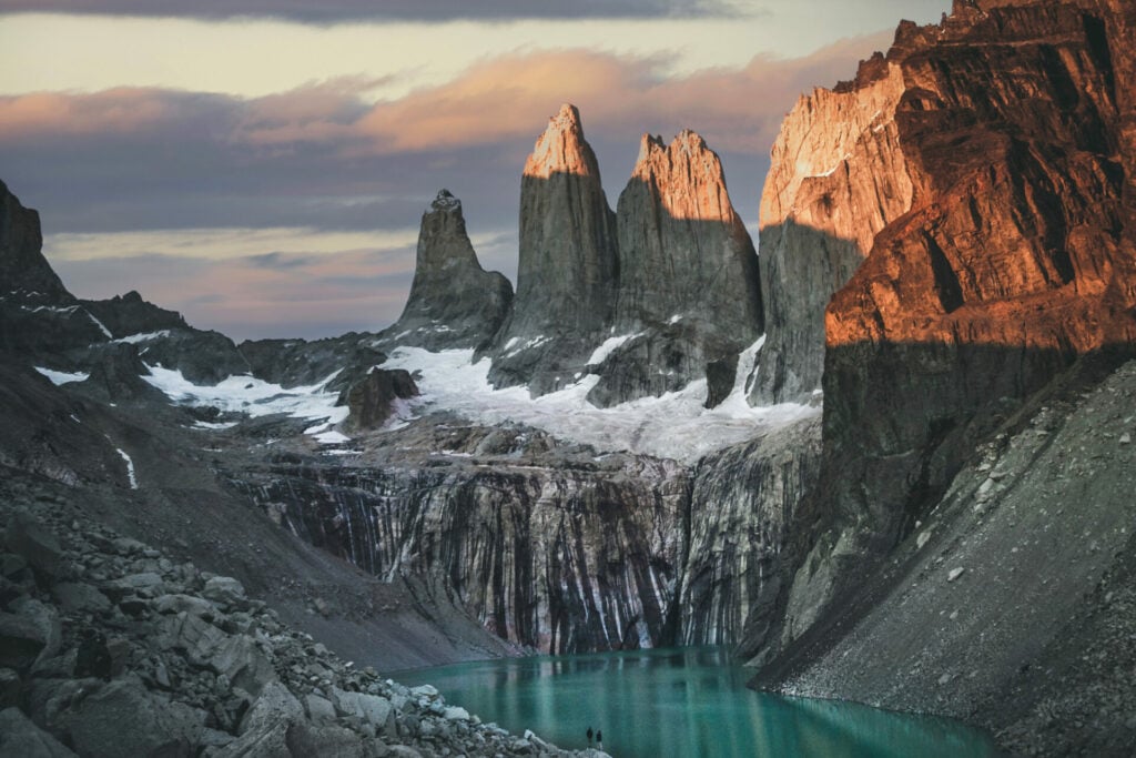 Au cœur de Voyages en Patagonie, trois grands pics rocheux se dressent au-dessus d'un lac turquoise, leurs pentes rocheuses étant saupoudrées de neige. La lumière du soleil éclaire certaines parties de la scène sous un ciel partiellement nuageux. - Karavaniers