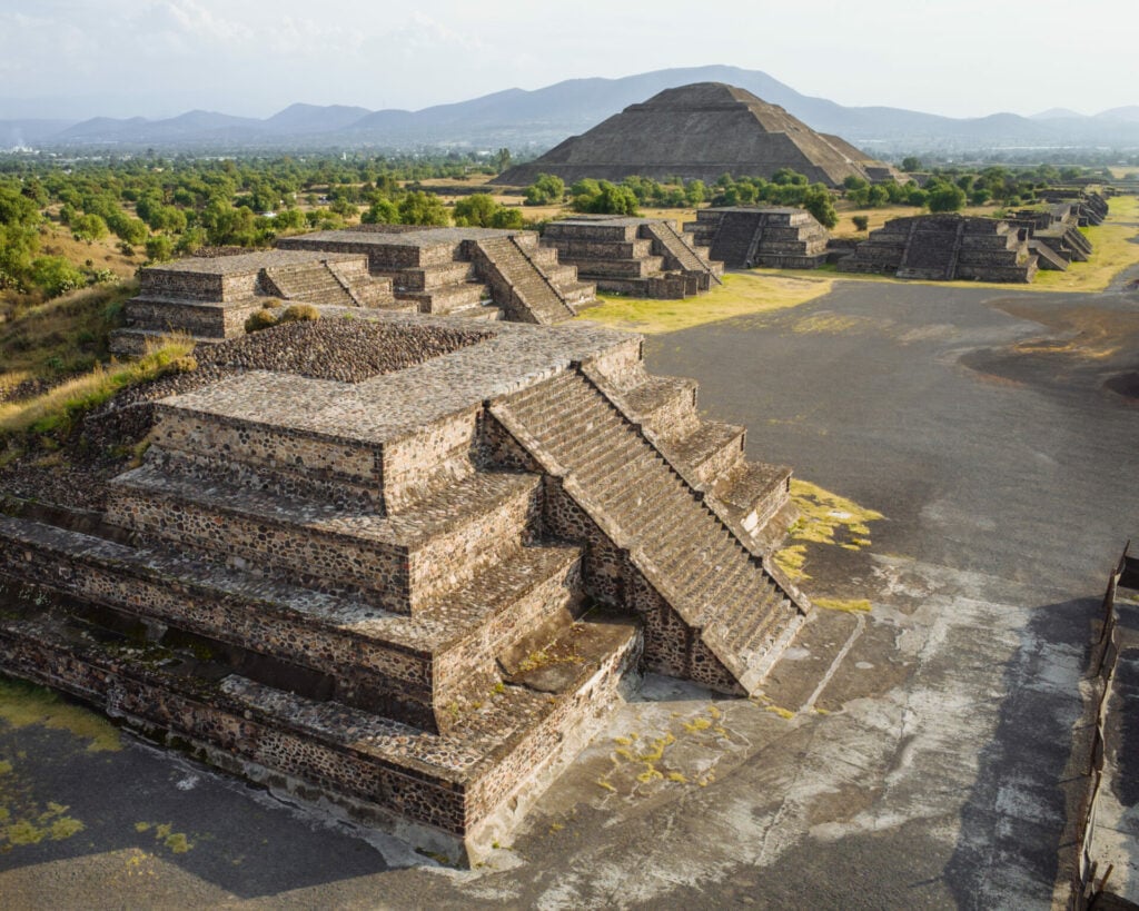 Des pyramides à degrés en pierre sont représentées à Teotihuacan, au Mexique, notamment la grande pyramide du Soleil, au milieu de la verdure et des montagnes lointaines, sous un ciel dégagé. La région fait partie des Volcans du Mexique et se trouve à proximité de plusieurs Pueblos Mágicos. - Karavaniers