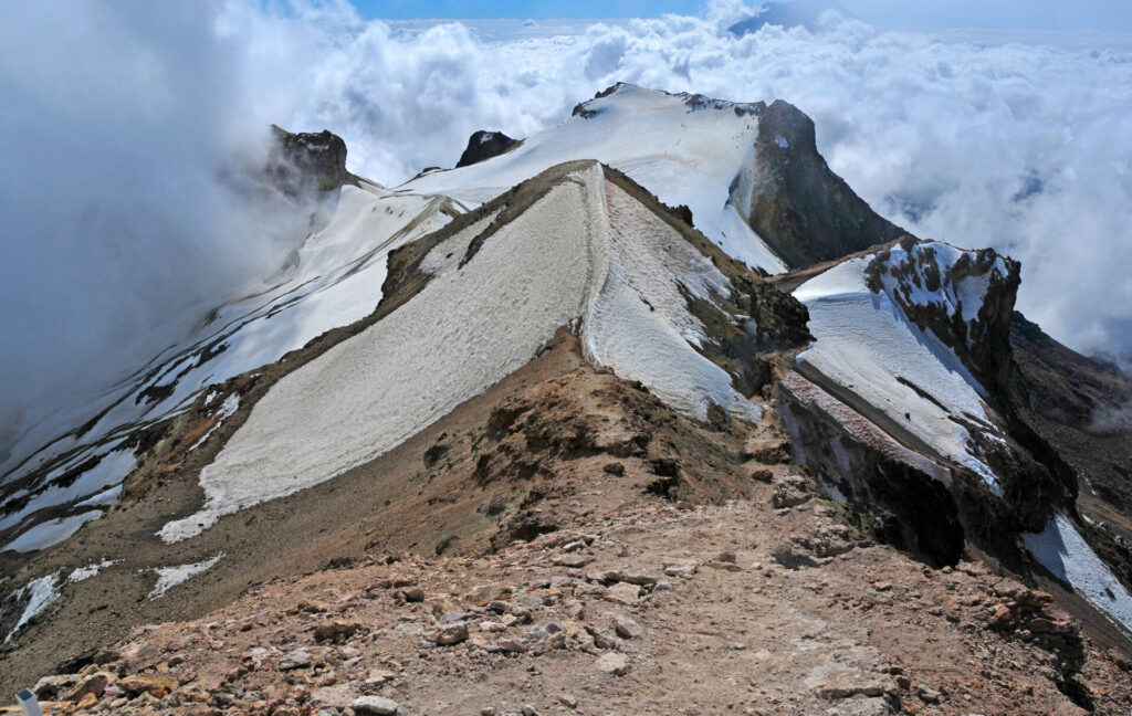 Une crête rocheuse avec des plaques de neige mène à des pics déchiquetés, rappelant les Volcans du Mexique, entourés de nuages et d'un ciel bleu. - Karavaniers