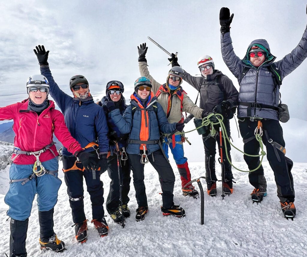 Sept Andinistes portant des vêtements d'hiver et du matériel d'escalade se tiennent sur une montagne enneigée, souriant et levant les bras en signe de célébration. Relié par des cordes, le groupe pose au sommet du Cayambe sous un ciel nuageux. - Karavaniers