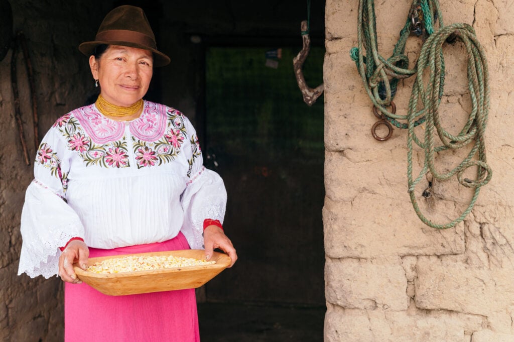 Une personne portant un chapeau marron, une blouse blanche brodée et une jupe rose se tient devant un mur d'adobe, tenant un plateau de grains de maïs - une scène peut-être familière aux Andinistes qui explorent la région près du Volcan Cayambe. Des cordes sont accrochées au mur à côté d'eux. - Karavaniers