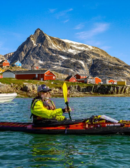 Kayakiste en tenue sèche naviguant dans un fjord, maisons rouges sur la côte