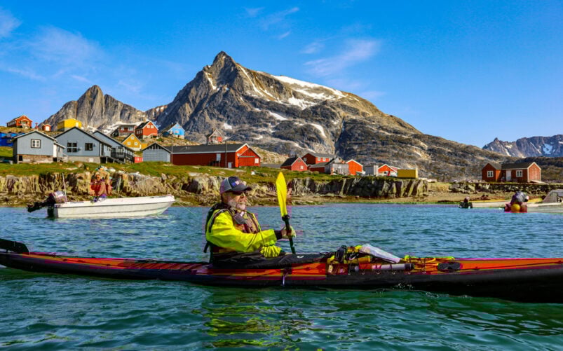 Kayakiste en tenue sèche naviguant dans un fjord, maisons rouges sur la côte