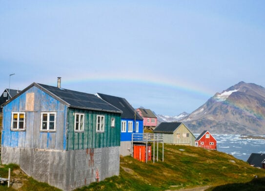Des maisons en bois colorées se dressent sur une colline herbeuse au bord d'une eau calme avec des glaces flottantes, tandis qu'un faible arc-en-ciel se dessine vers un pic montagneux - une scène idéale pour explorer les Fjords de l'est en kayak et trek. - Karavaniers