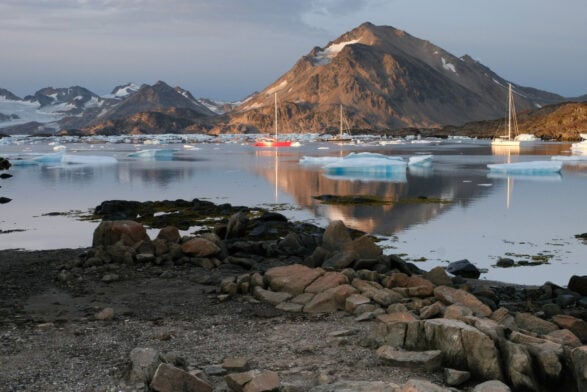Rivage rocheux avec des icebergs épars flottant dans une eau calme ; trois voiliers, un rouge et deux blancs, sont ancrés au milieu des majestueux Fjords de l'est en kayak et trek, avec des montagnes enneigées s'élevant sous un ciel nuageux. - Karavaniers