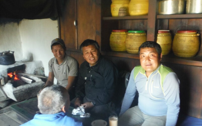 Scène conviviale à l’intérieur d’une maison népalaise, où quatre hommes partagent un moment autour d’un poêle à bois, entourés de pots traditionnels en argile.