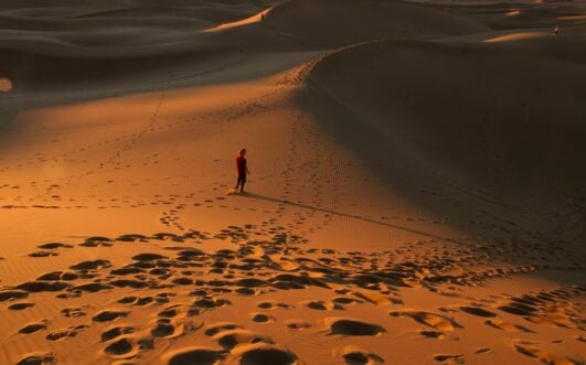 Une personne vêtue de rouge marche seule sur des dunes de sable au coucher du soleil, projetant une longue ombre. De nombreuses empreintes de pas sont visibles dans le sable doré, et des ondulations créent des motifs texturés à la surface. - Karavaniers
