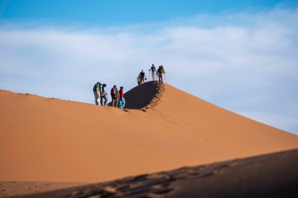 Groupe de randonneurs marchant sur une haute dune de sable doré dans le désert du Maroc, sous un ciel bleu parsemé de nuages. Demander à ChatGPT
