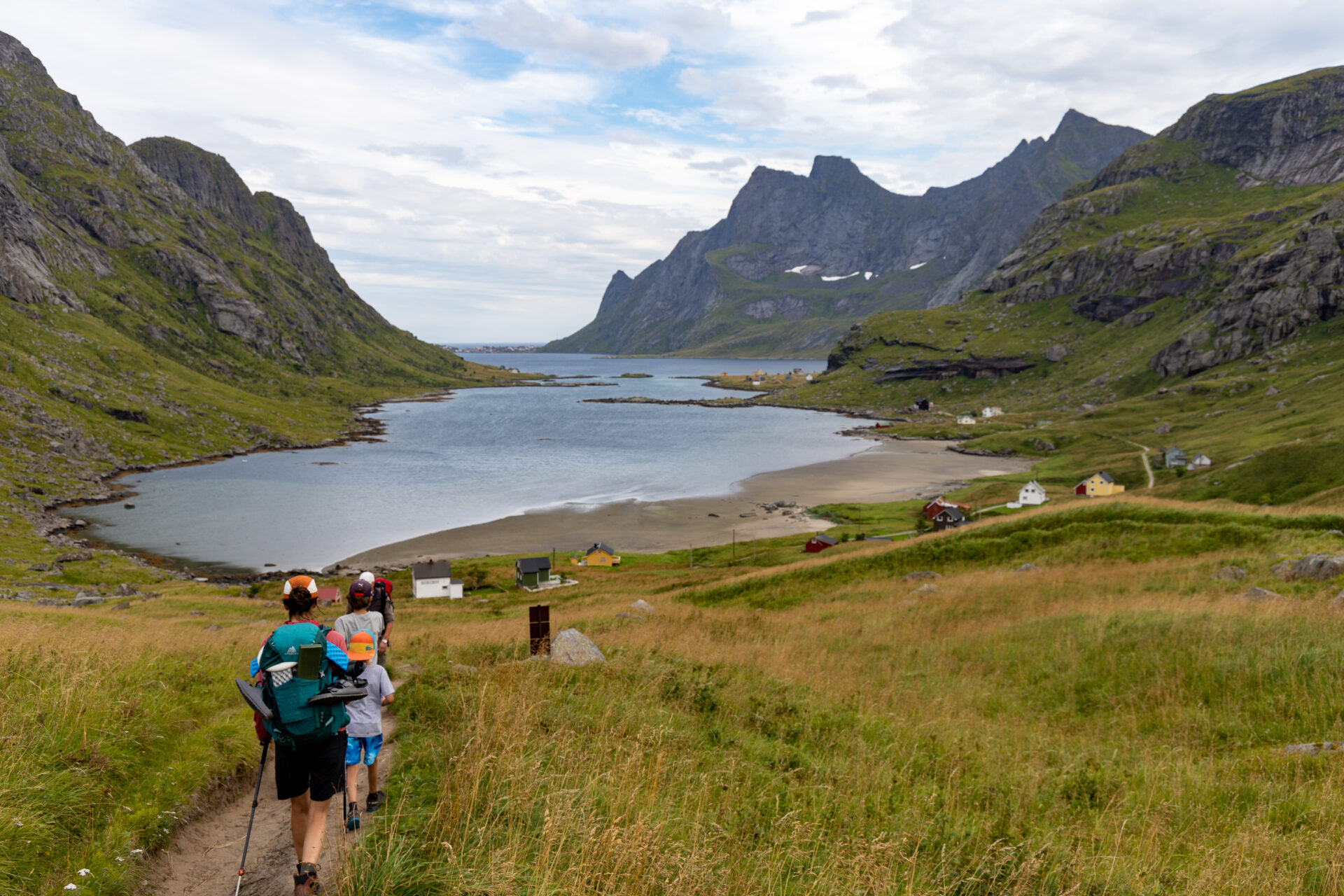 Famille en randonnée dans les îles Lofoten, chemin herbeux menant vers un fjord entouré de montagnes abruptes en Norvège.