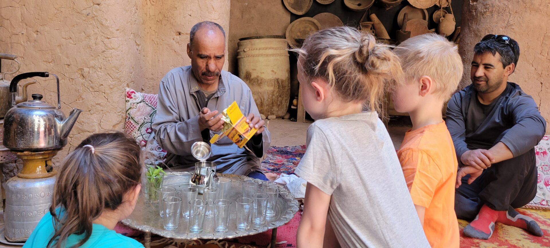 Un homme prépare le thé à la menthe traditionnel devant un groupe d’enfants attentifs, dans une maison en terre du sud marocain, au décor simple et chaleureux