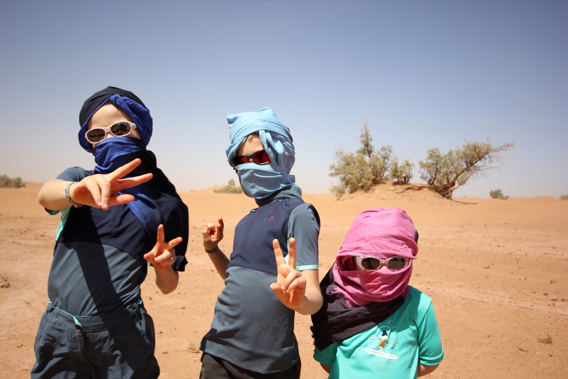 Trois enfants vêtus de chèches et de lunettes de soleil posent en souriant dans le désert marocain, sur fond de sable doré et de quelques arbres clairsemés