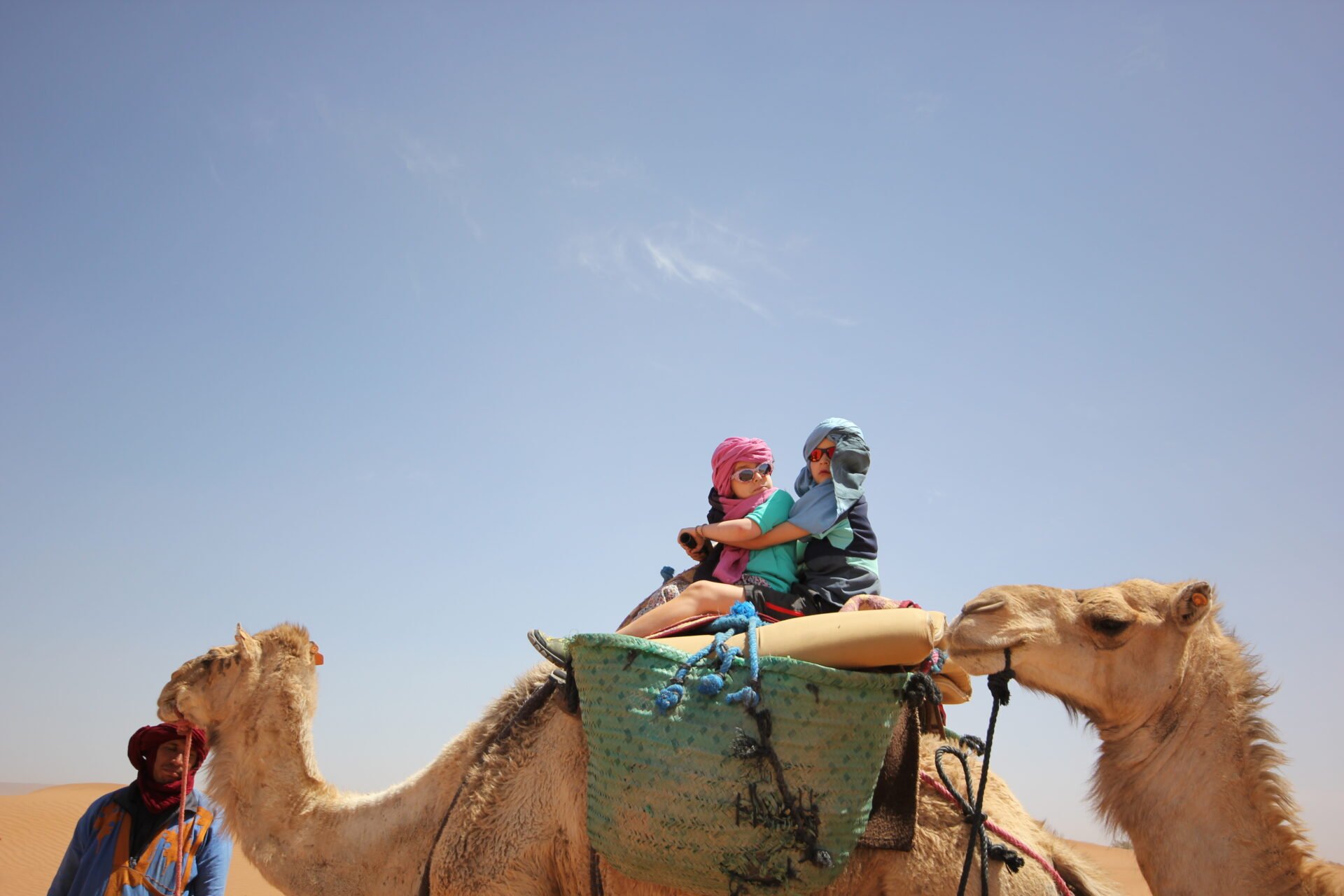 Deux enfants aux foulards colorés montés sur un dromadaire dans le désert marocain, accompagnés d’un chamelier, sous un ciel dégagé