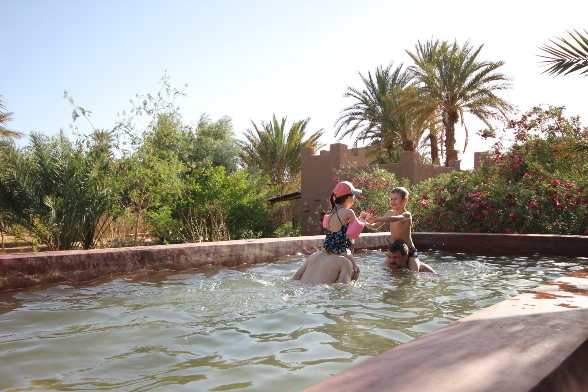 Deux enfants se rafraîchissent dans une piscine entourée de palmiers et de végétation luxuriante, dans une oasis au sud du Maroc