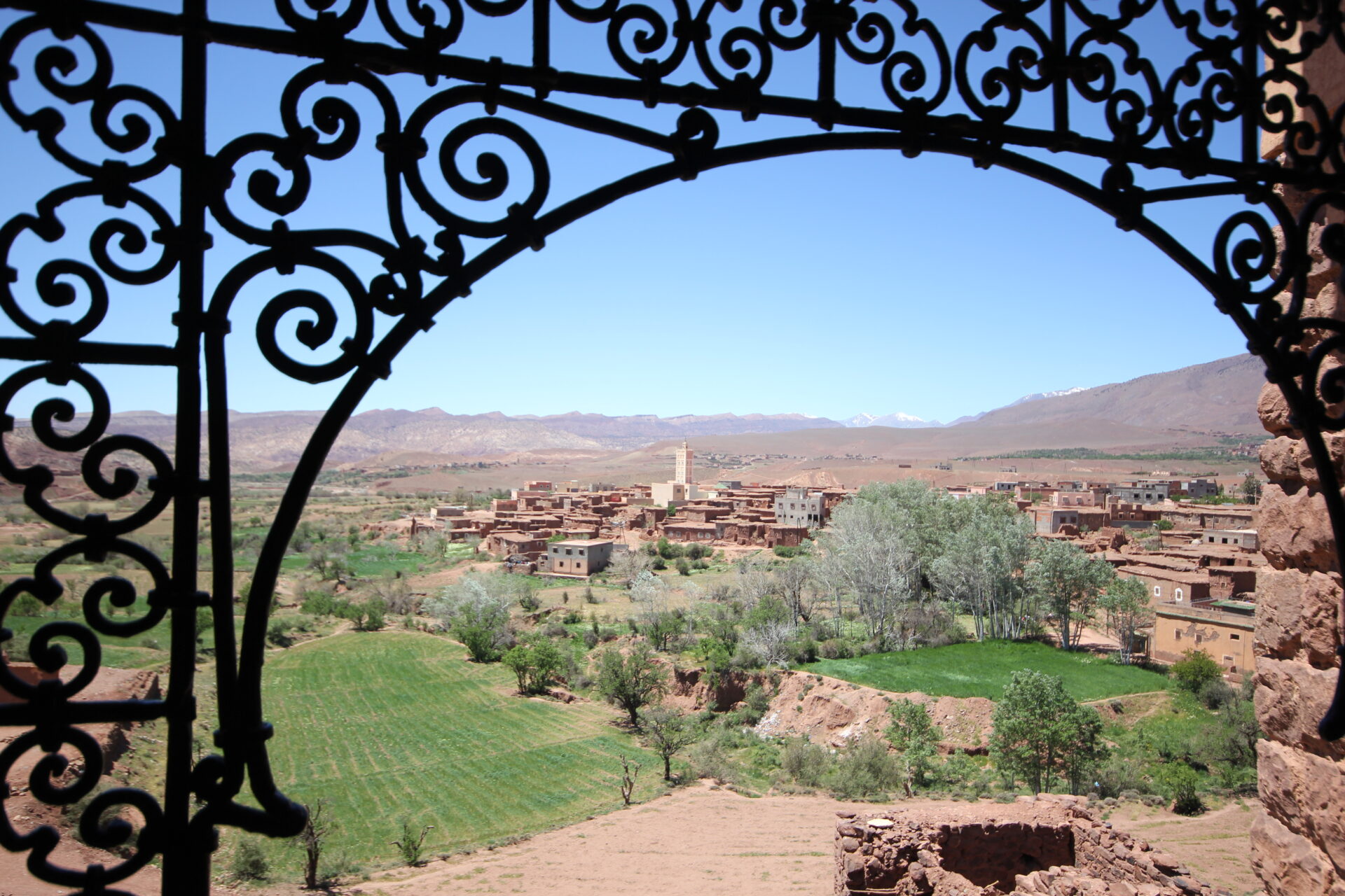 Vue encadrée par une arche en fer forgé sur un village marocain en pisé, entouré d’oasis et de champs verdoyants, avec les montagnes en toile de fond
