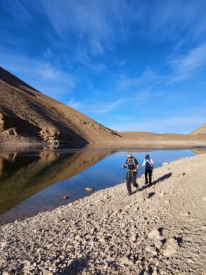 Deux randonneurs au bord d’un lac de montagne entouré de collines arides dans la région de Tessaout, Maroc.