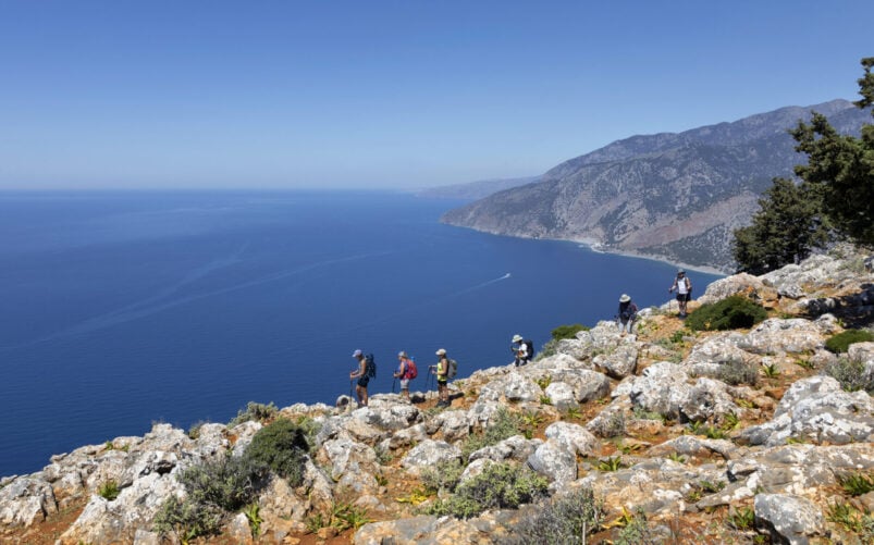 Groupe de randonneurs longeant un sentier côtier rocailleux avec vue panoramique sur la mer Égée en contrebas