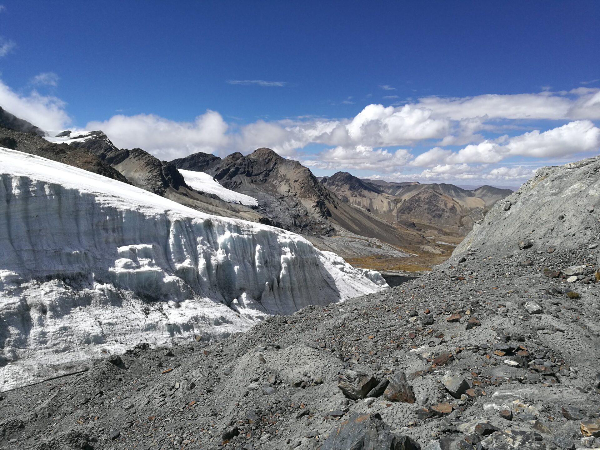 Vue d’un glacier abrupt dans la Cordillère Royale en Bolivie, avec des parois de glace blanche contrastant avec les roches grises environnantes, sous un ciel bleu parsemé de nuages.