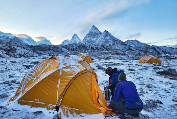 Deux personnes sont agenouillées à côté d'une tente jaune dans un paysage enneigé et rocheux - une scène typique d'un voyage de longue durée - tandis que d'autres tentes jaunes parsèment la zone et que des montagnes enneigées s'élèvent sous un ciel nuageux. - Karavaniers