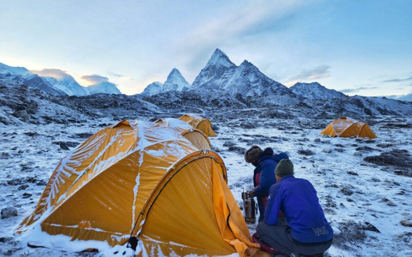 Deux personnes sont agenouillées à côté d'une tente jaune dans un paysage enneigé et rocheux - une scène typique d'un voyage de longue durée - tandis que d'autres tentes jaunes parsèment la zone et que des montagnes enneigées s'élèvent sous un ciel nuageux. - Karavaniers