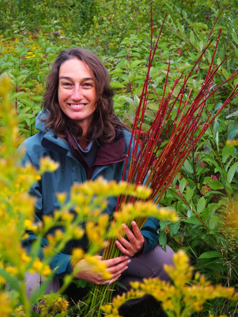 Malie Lessard Therrien, guide chez Karavaniers, souriante assise dans un champ de fleurs jaunes, tenant une gerbe de branches rouges, entourée d’une végétation verdoyante.