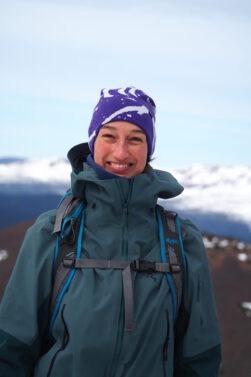 Malie Lessard Therrien, guide chez Karavaniers, souriante en habit de randonnée et tuque mauve, debout en montagne avec un paysage de sommets enneigés à l’arrière-plan.