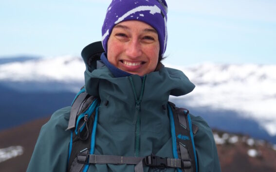 Malie Lessard Therrien, guide chez Karavaniers, souriante en habit de randonnée et tuque mauve, debout en montagne avec un paysage de sommets enneigés à l’arrière-plan.