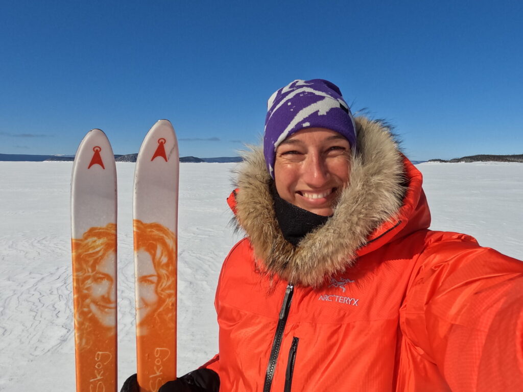Malie Lessard Therrien, guide chez Karavaniers, souriante en manteau d’expédition orange avec capuchon bordé de fourrure, tenant des skis sur un lac gelé sous un ciel bleu éclatant.