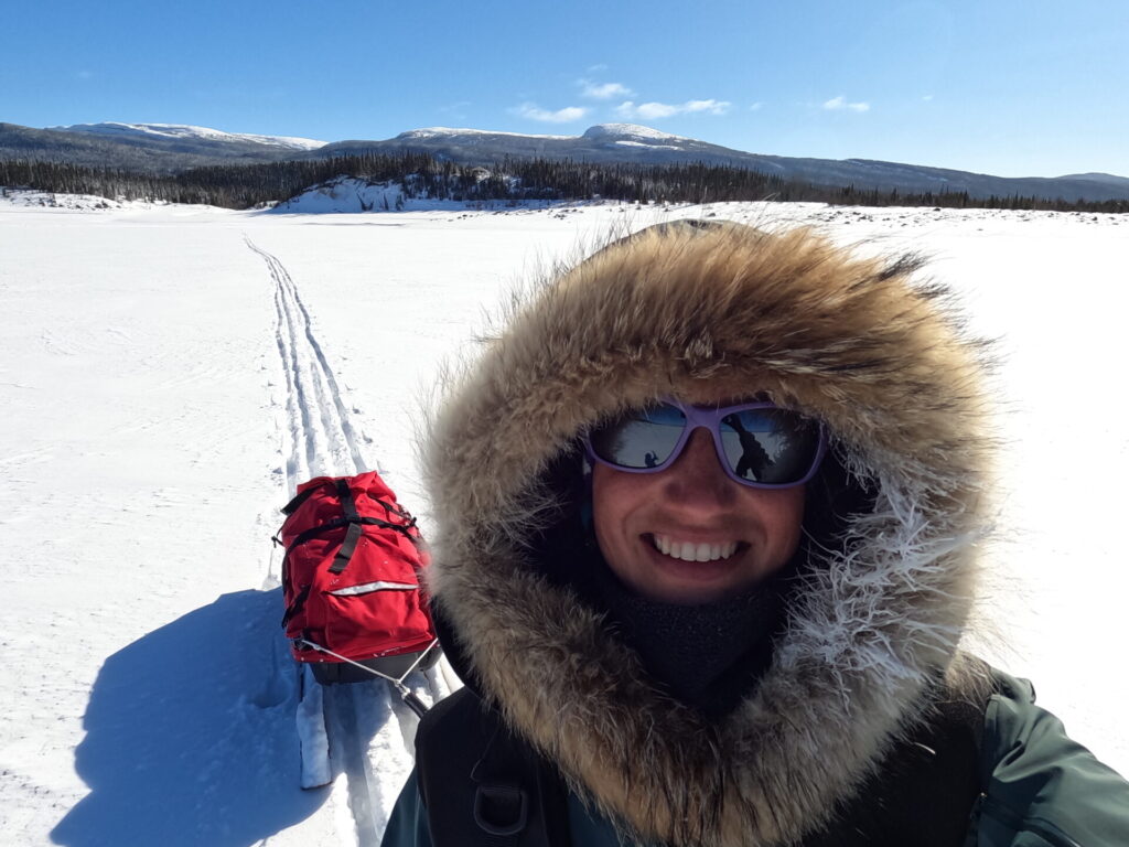 Malie Lessard Therrien, guide chez Karavaniers, souriante en manteau d’expédition avec capuchon bordé de fourrure, tirant un traîneau rouge sur une étendue enneigée sous un ciel bleu.