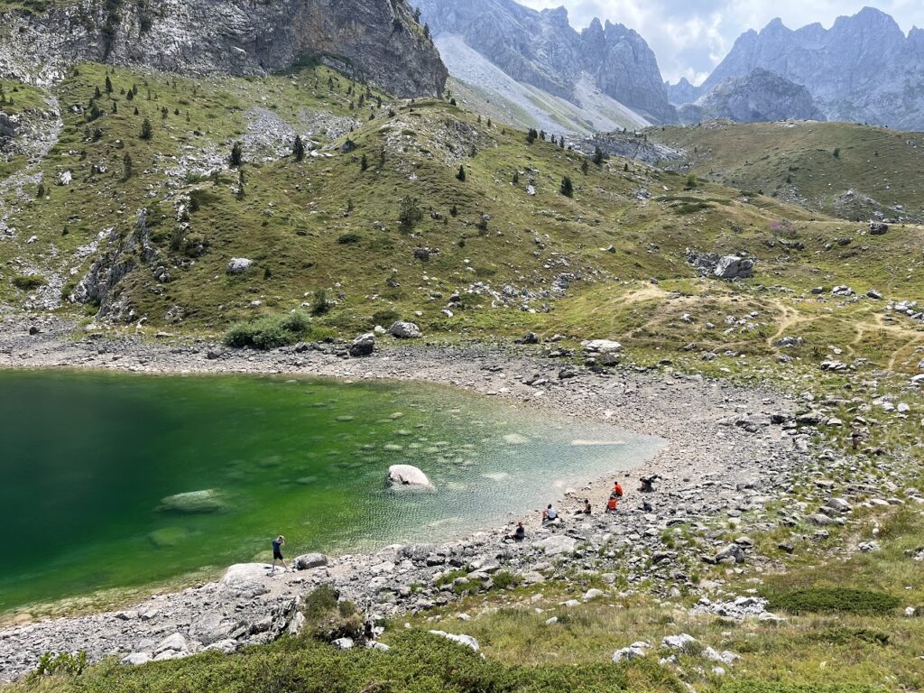 Un petit groupe de personnes se tient debout et assis sur la rive rocheuse d'un lac vert dans les Alpes Dinariques, entouré de collines herbeuses et de montagnes lointaines sous un ciel partiellement nuageux. - Karavaniers