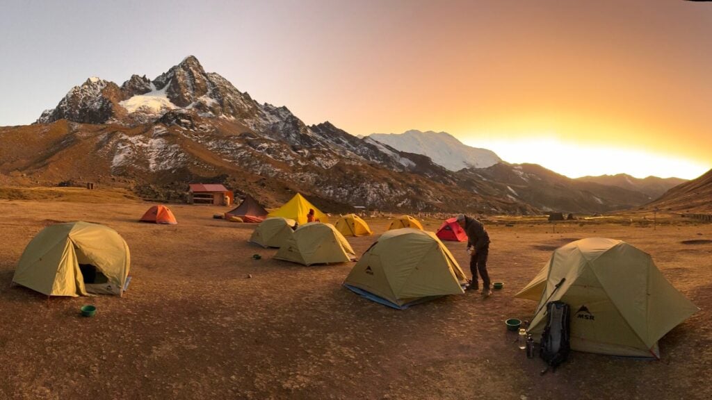 Plusieurs tentes jaunes et orange sont installées sur un sol sec au pied de l'Ausangate enneigé pendant le lever ou le coucher du soleil. Une personne se tient près d'une tente, tandis que la lumière chaude du soleil et un ciel clair illuminent cette majestueuse scène de montagne. - Karavaniers