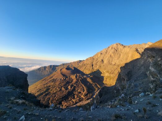 Paysage de montagnes rocheuses au lever du soleil, avec la lumière du soleil illuminant les pentes et un cratère près du centre - rappelant le Kilimandjaro. Le ciel est clair et bleu, avec des nuages lointains visibles sur la gauche. Un cadre parfait pour une conférence sur les merveilles de la nature. - Karavaniers