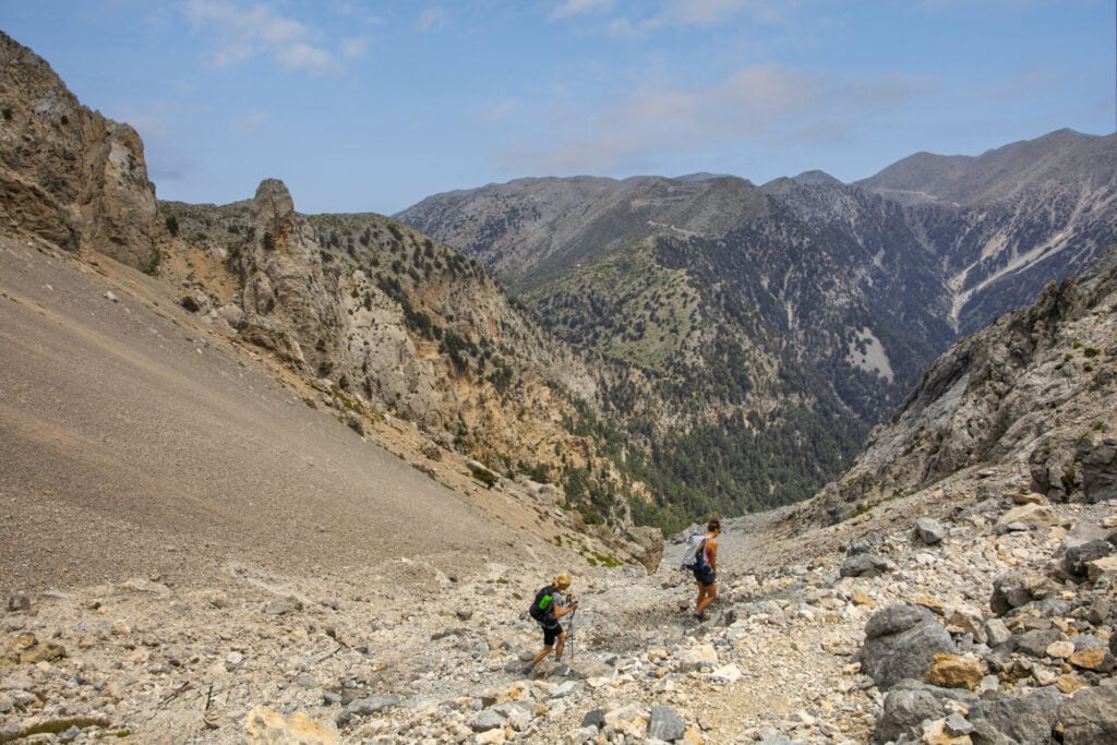 Deux randonneurs avec des sacs à dos marchent sur une pente rocheuse entourée de pics escarpés, capturant l'esprit des voyages sur mesure sous un ciel partiellement nuageux. - Karavaniers