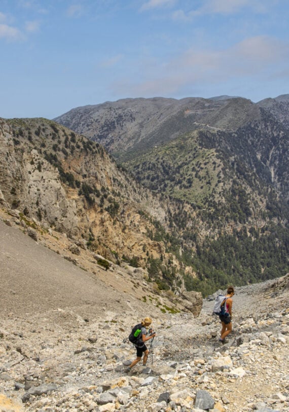 Deux randonneurs avec des sacs à dos marchent sur une pente rocheuse entourée de pics escarpés, capturant l'esprit des voyages sur mesure sous un ciel partiellement nuageux. - Karavaniers