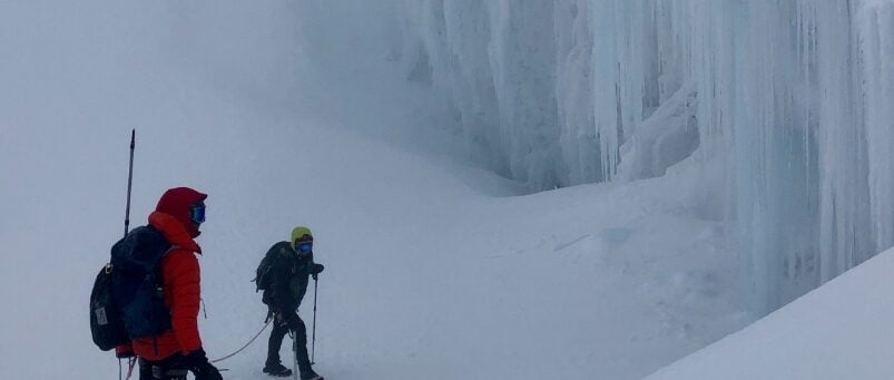 Deux personnes en tenue d'hiver, avec sac à dos et bâtons de ski, se tiennent sur une pente enneigée près d'un mur de grandes stalactites, entourées de brouillard - une aventure digne des Karavaniers des hautes montagnes. - Karavaniers