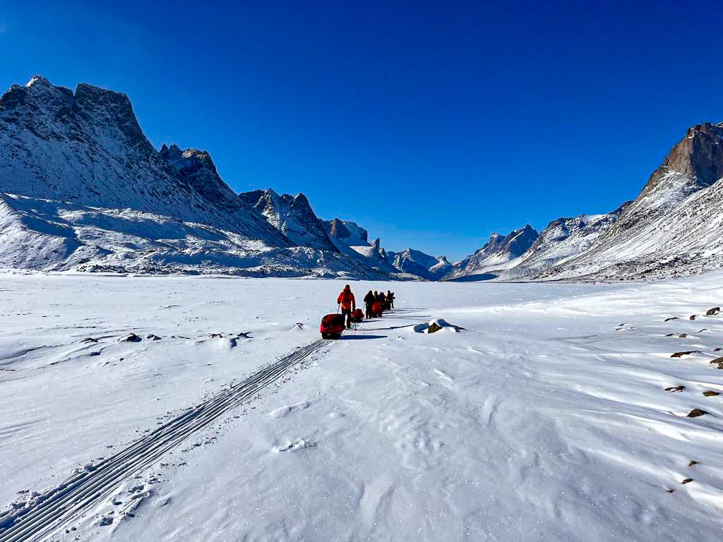 Un groupe de personnes en tenue d'hiver tire des traîneaux sur un paysage enneigé et plat de la Terre de Baffin, entouré de montagnes escarpées couvertes de glaces éternelles, sous un ciel bleu limpide. - Karavaniers