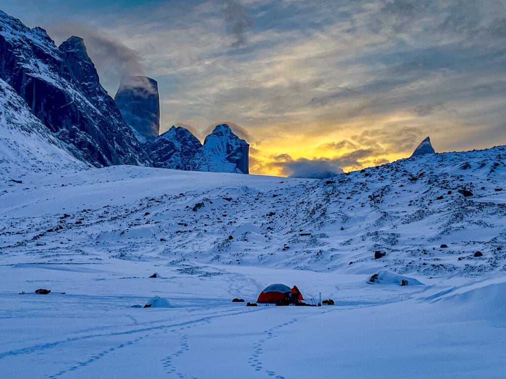 Une seule tente orange se dresse sur le paysage montagneux et enneigé de l'expédition à Baffin au lever du soleil. Des pics déchiquetés se dressent à l'arrière-plan, des empreintes de pas sur les glaces éternelles et une lueur dorée illuminent le ciel partiellement nuageux. - Karavaniers