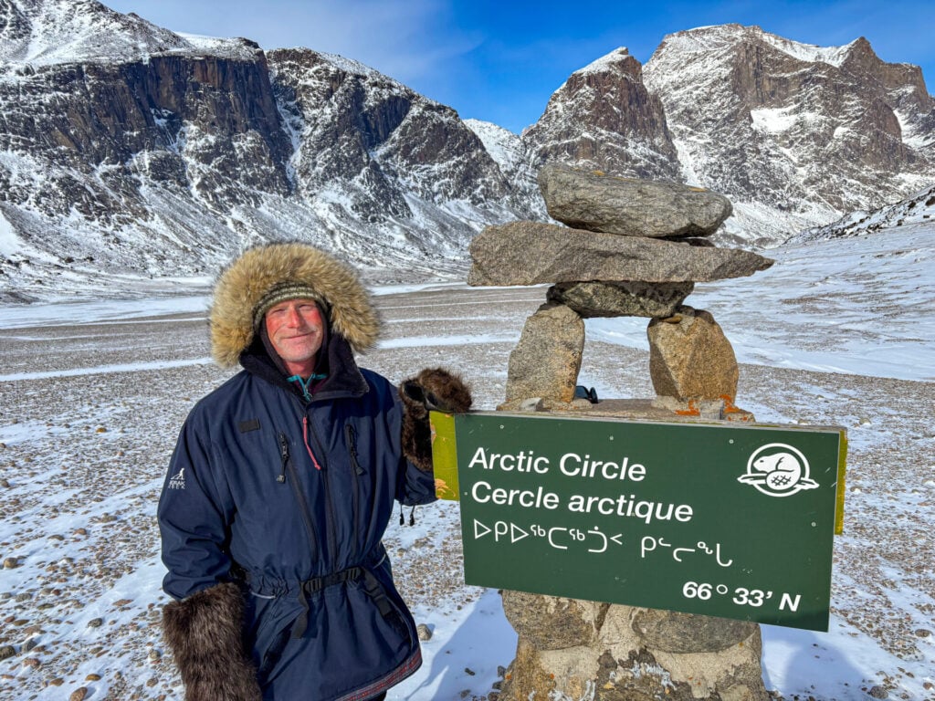 Une personne vêtue d'une parka doublée de fourrure se tient à côté d'un cairn de pierre et d'un panneau vert marquant le cercle polaire arctique à 66°33′N, entouré de glaces éternelles et de montagnes escarpées - une scène évocatrice de l'Expédition à Baffin, Pays des glaces. - Karavaniers