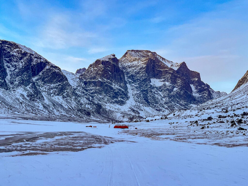 Les montagnes enneigées sous un ciel partiellement nuageux s'élèvent au-dessus des glaces éternelles de Baffin. Dans le premier plan plat et gelé, plusieurs tentes orange d'une expédition se détachent vivement sur la neige près du centre de l'image. - Karavaniers