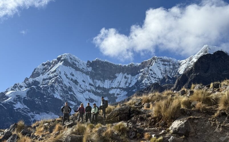 Cinq personnes se trouvent sur une pente rocheuse et herbeuse près d'Ausangate, avec des montagnes enneigées s'élevant derrière eux sous un ciel bleu avec des nuages. - Karavaniers