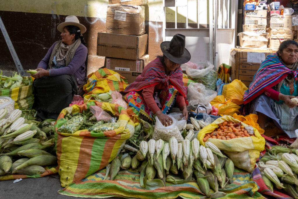 Trois femmes sont assises sur le sol d'un marché, entourées de grands sacs de maïs et de haricots, comme dans les scènes du Trek du Huayhuash. Derrière elles, des boîtes en carton sont empilées et elles portent des chapeaux et des vêtements colorés. - Karavaniers