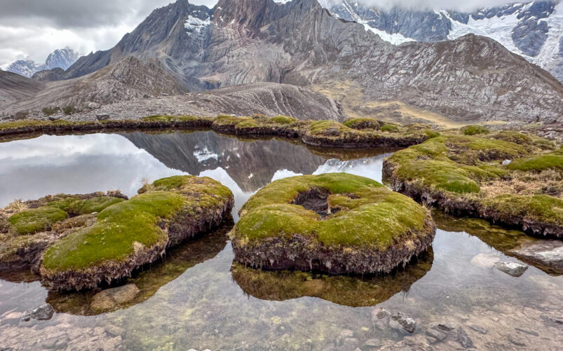 Un lac d'altitude situé le long du Trek du Huayhuash présente de petites îles rondes et herbeuses flottant sur l'eau calme, reflétant des montagnes rocheuses avec des taches de neige sous un ciel nuageux. - Karavaniers